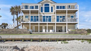 Una casa fronte spiaggia a più piani con balconi, un portico coperto e una spiaggia sabbiosa in primo piano.