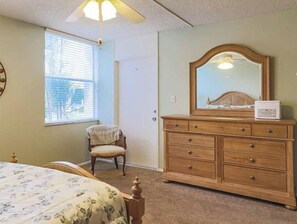 A bedroom with a bed, a wooden dresser, a chair, a window with blinds, and a ceiling fan.