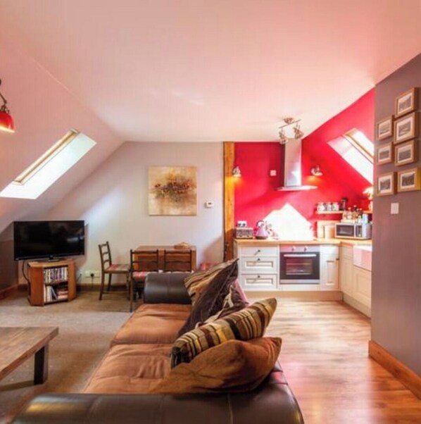 Ein gemütliches Wohnzimmer mit einer Couch und einem Fernseher, eine Küche mit roten Wänden sowie eine Essecke mit Tisch und Stühlen.