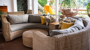 Un canapé en osier avec des coussins, un fauteuil suspendu et une petite table, le tout dans un espace intérieur bien éclairé, avec vue sur la verdure à l’extérieur.