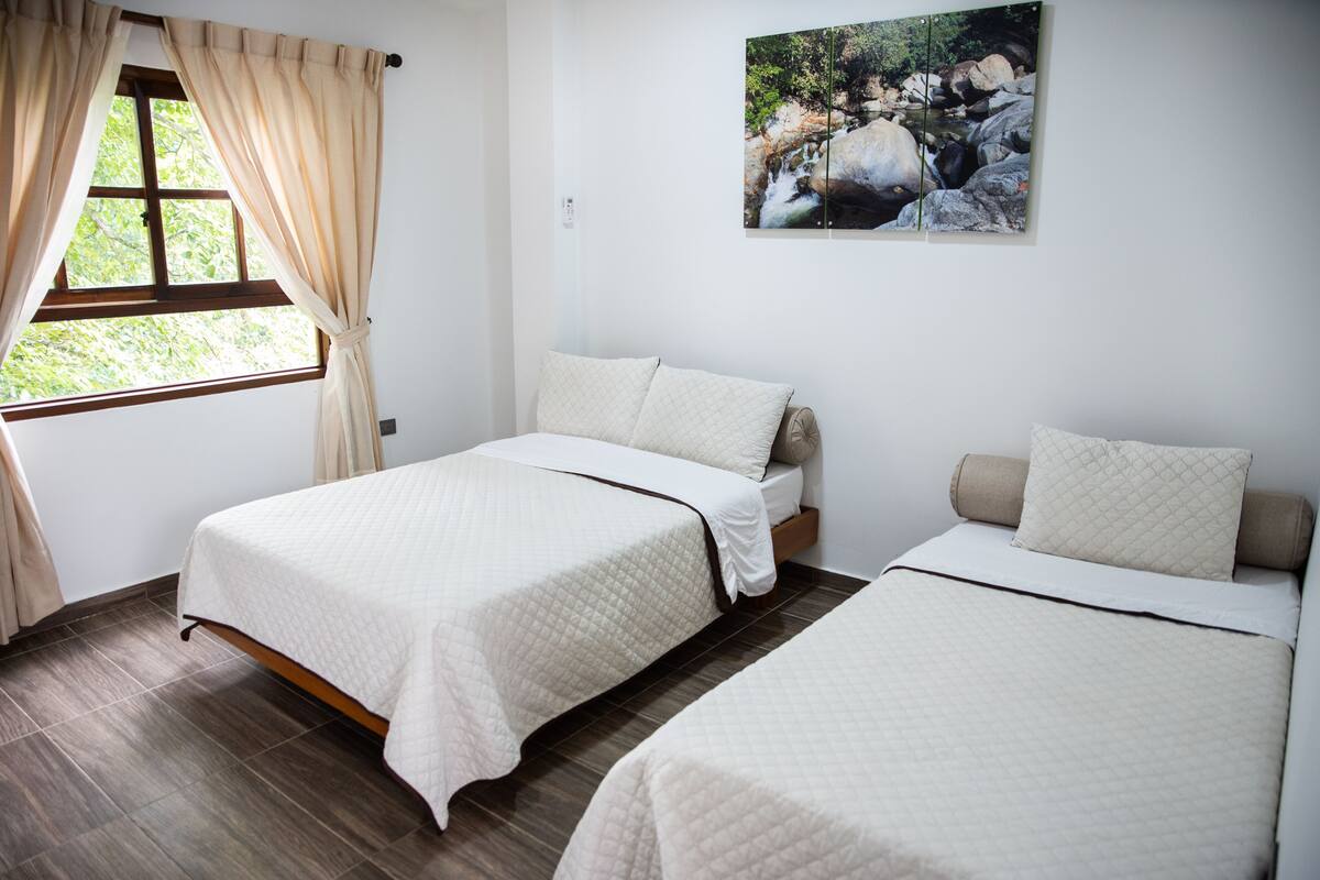 A room with two beds, a window with curtains, and a wall-mounted picture of a natural scene.