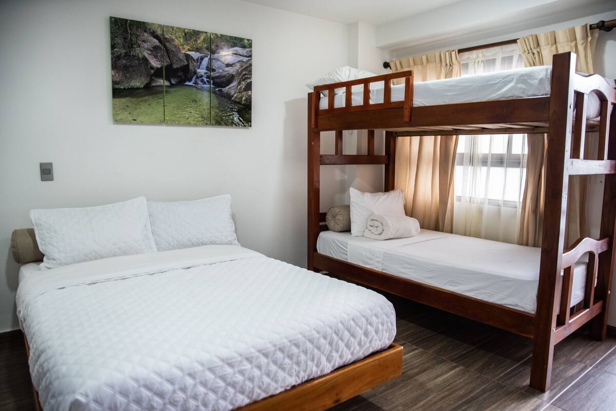 A bedroom with a bunk bed and a single bed, both with white bedding. There is a painting of a natural scene on the wall.