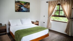 Une chambre avec un lit en bois, un plaid vert, une table de chevet, une lampe de chevet et une fenêtre avec des rideaux.