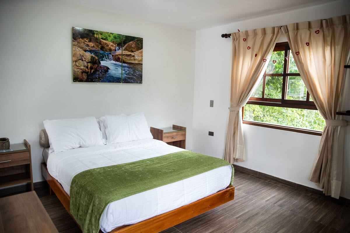 A bedroom with a wooden bed, a green throw blanket, a nightstand, a bedside table, and a window with curtains.