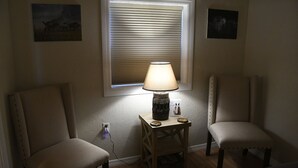 Ein Zimmer mit zwei Stühlen, einem Tisch mit Lampe, einem Fenster mit Jalousien und einer Wandsteckdose.