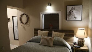Ein Schlafzimmer mit Bett, Nachttisch mit Lampe, Wanduhr, Fenster mit Jalousien, einer Tür und einem gerahmten Bild an der Wand.