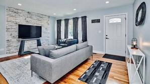 A modern living room with a grey sofa, a flat-screen TV on a wooden stand, and a white door with a 'FAMILY' sign. - Fox Valley Farmhouse by the river - walk to town & pool (St. Charles)