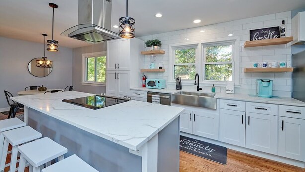 A modern kitchen with white cabinets, a central island, pendant lighting, and a view of greenery outside. - Fox Valley Farmhouse by the river - walk to town & pool (St. Charles)
