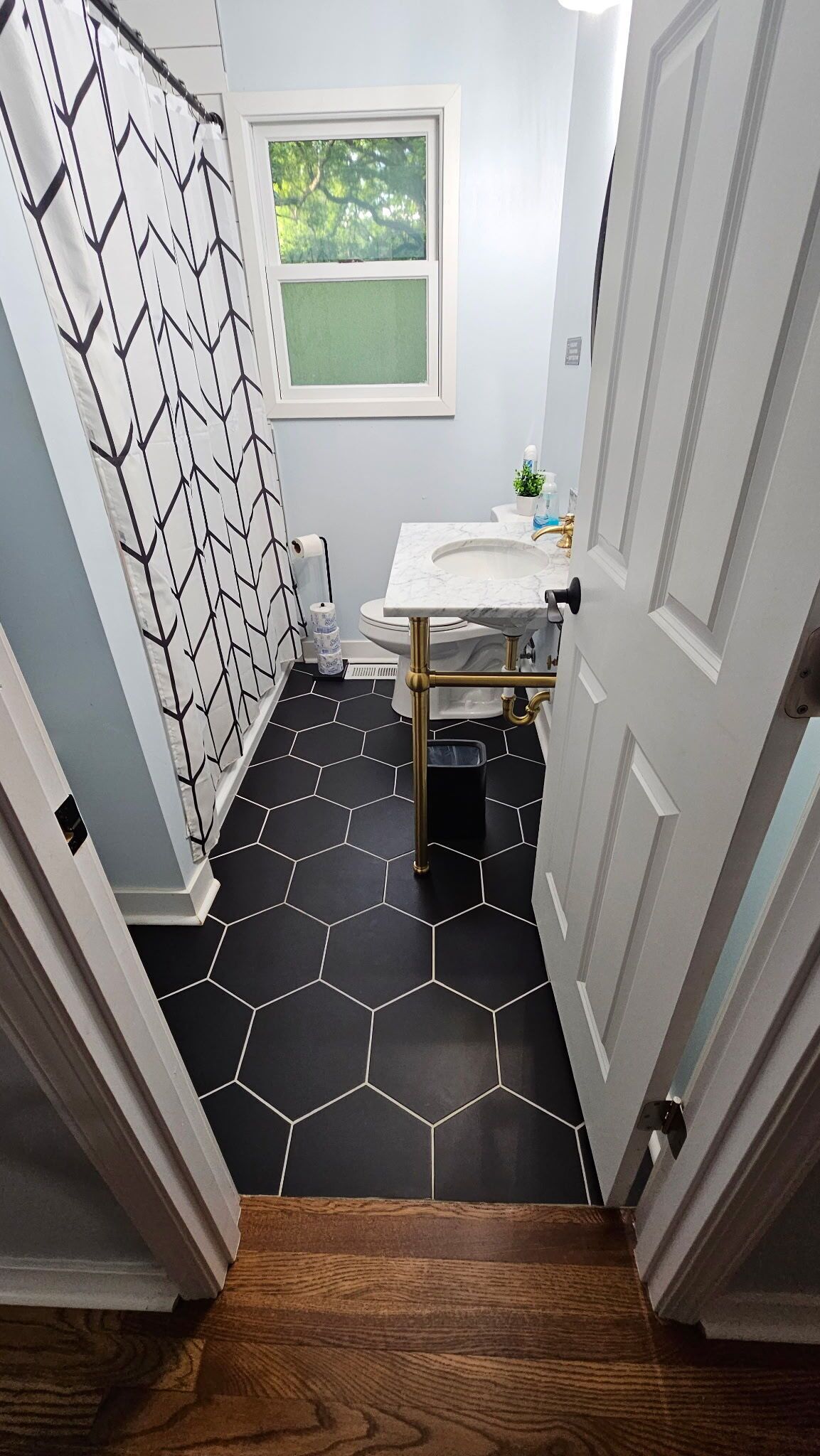 A bathroom with a white sink, black hexagonal tiles, a shower curtain with a geometric pattern, and a window with a view of trees.