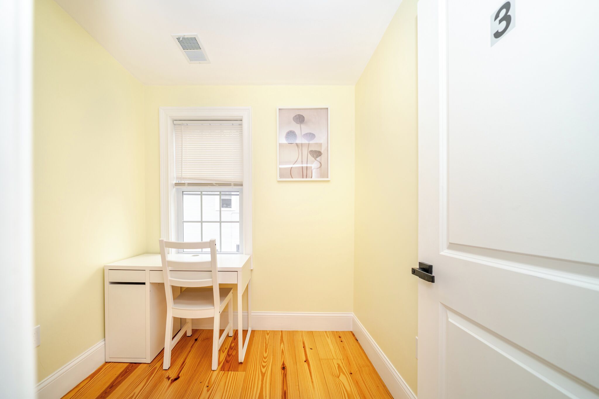 A small, bright room with a white desk and chair, a window with blinds, and a door with a number.
