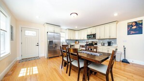 A modern kitchen with white cabinets, stainless steel appliances, and a dining area with a wooden table and chairs. - Spacious Greater Boston Townhouse Near I-93 • Parking • Pet-Friendly | 37 (Quincy)
