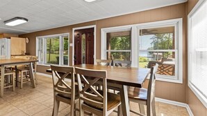Un coin repas avec une table et des chaises en bois, une cuisine équipée d’un réfrigérateur et de grandes fenêtres avec des stores.
