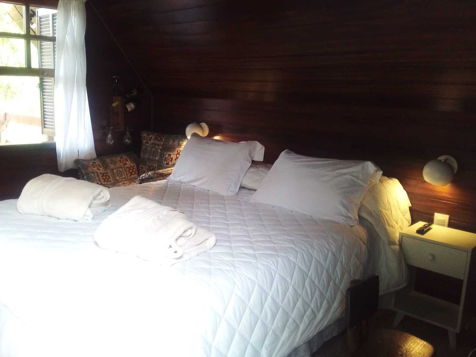 A cozy, dimly lit room with a wooden ceiling and walls, featuring a neatly made bed with white linens and pillows, a small bedside table with a lamp, and a patterned armchair.