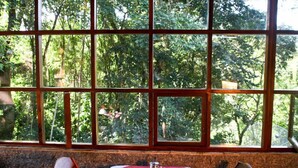 Une table à manger dressée pour deux personnes, recouverte d’une nappe rouge et blanche, placée devant une grande fenêtre donnant sur un paysage arboré.