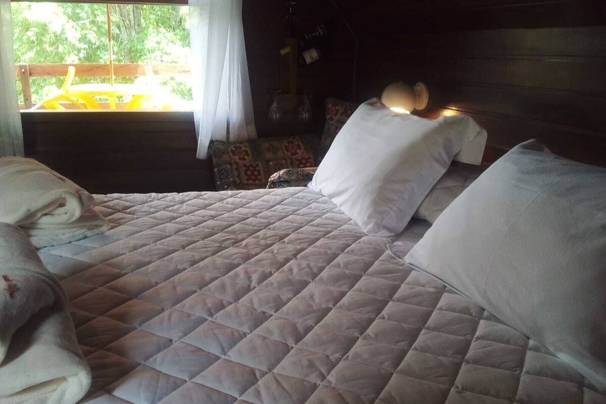Ein Bett mit einer gesteppten Decke, ein Nachttisch mit einer Lampe und Blick ins Grüne.