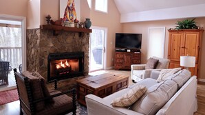 A cozy living room with a stone fireplace, comfortable seating, and a television. - Witt's End at Mill Creek: Walk to Downtown | Creekside Retreat w/ Screened Porch (Highlands)