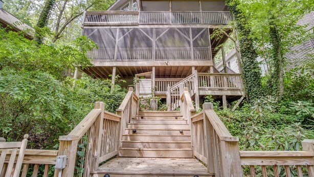 A wooden deck leading to a multi-level wooden house surrounded by dense foliage. - Witt's End at Mill Creek: Walk to Downtown | Creekside Retreat w/ Screened Porch (Highlands)