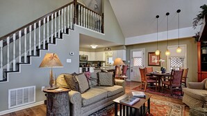 A spacious living area with a staircase, a dining table with chairs, and a kitchen in the background. - Heinrich - Mill Creek Cottage: Walk to Downtown | Creekside Retreat w/ Hot Tub (Highlands)