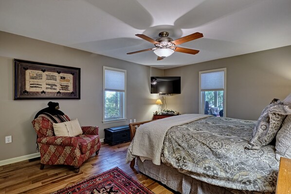 A bedroom with a large bed, a ceiling fan, a framed picture on the wall, a chair, and a TV mounted on the wall. - Heinrich - Mill Creek Cottage: Walk to Downtown | Creekside Retreat w/ Hot Tub (Highlands)