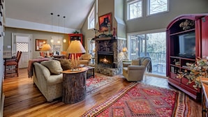 A spacious living room with a stone fireplace, wooden flooring, and large windows. - Heinrich - Mill Creek Cottage: Walk to Downtown | Creekside Retreat w/ Hot Tub (Highlands)