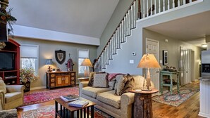 A spacious living room with a high ceiling, a staircase, and a cozy fireplace. - Heinrich - Mill Creek Cottage: Walk to Downtown | Creekside Retreat w/ Hot Tub (Highlands)