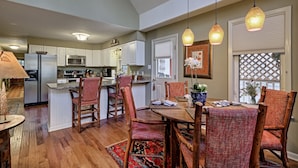 A spacious kitchen with white cabinets, a central island, and a dining area with wooden chairs and a table. - Heinrich - Mill Creek Cottage: Walk to Downtown | Creekside Retreat w/ Hot Tub (Highlands)