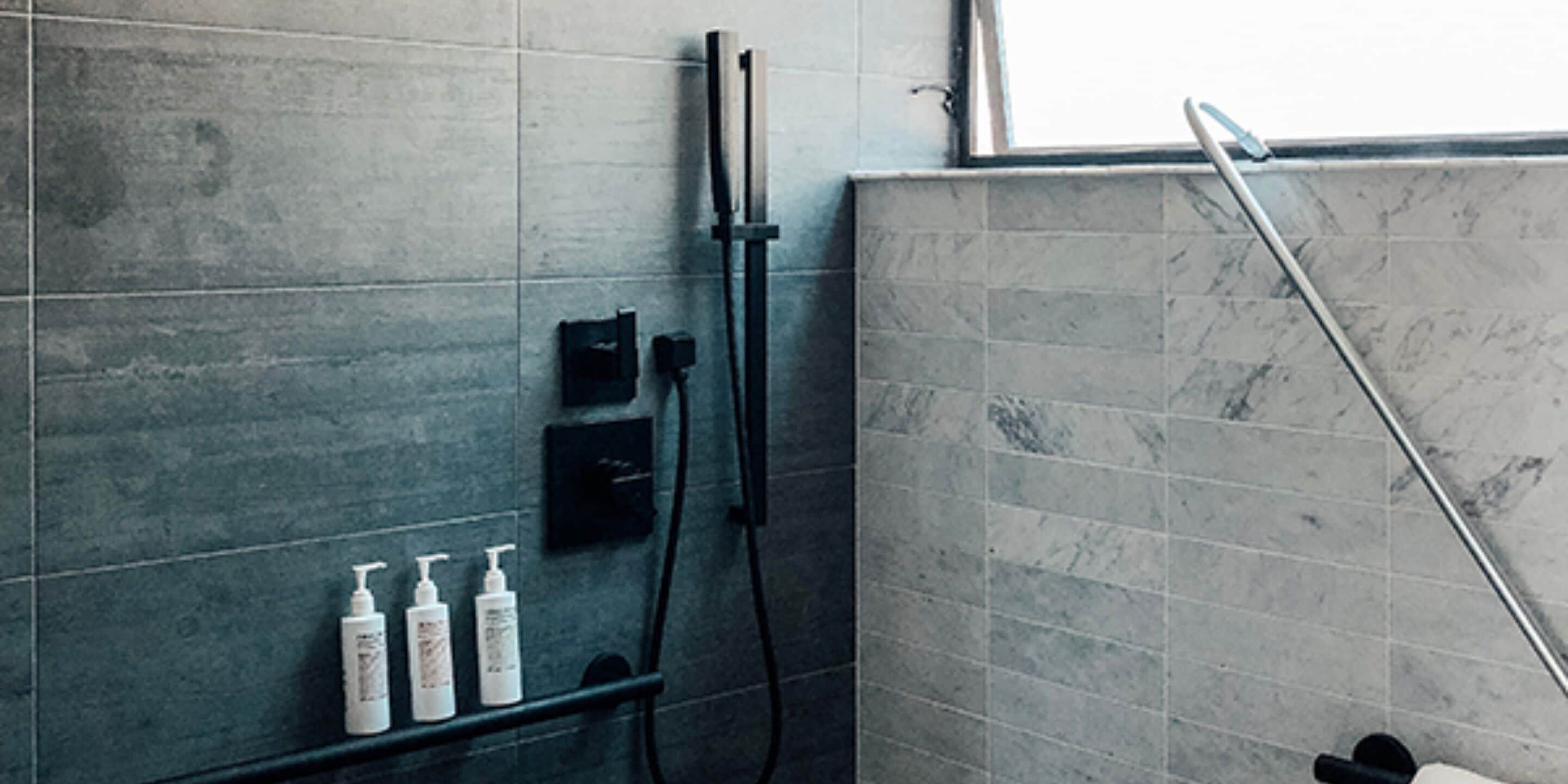 A bathroom with grey tiled walls, a shower area, and a shelf with three bottles of toiletries.