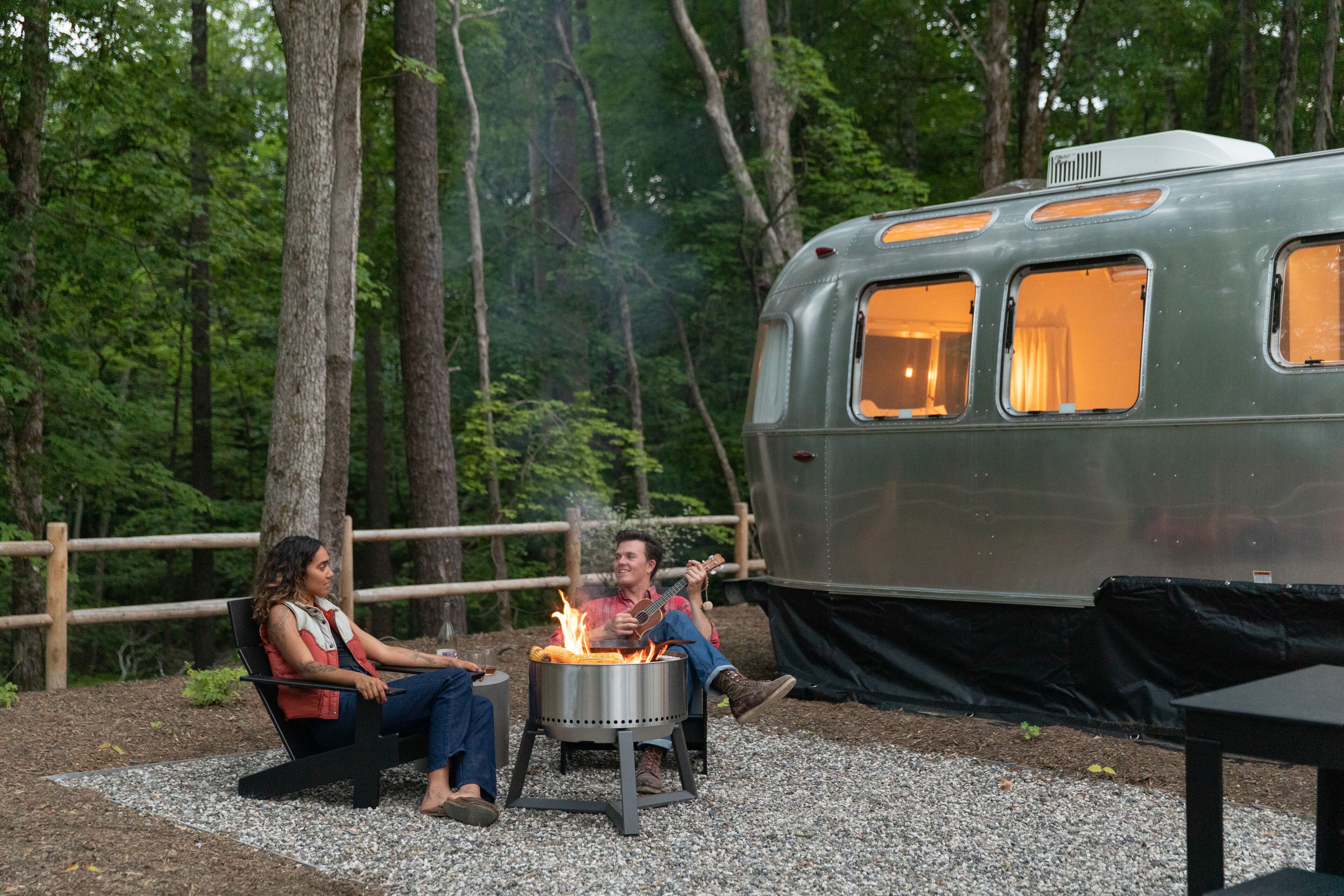 A couple sitting by a campfire with a vintage camper parked nearby.