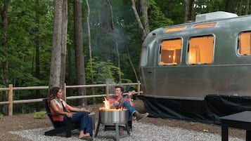 A couple sitting by a campfire with a vintage camper parked nearby.