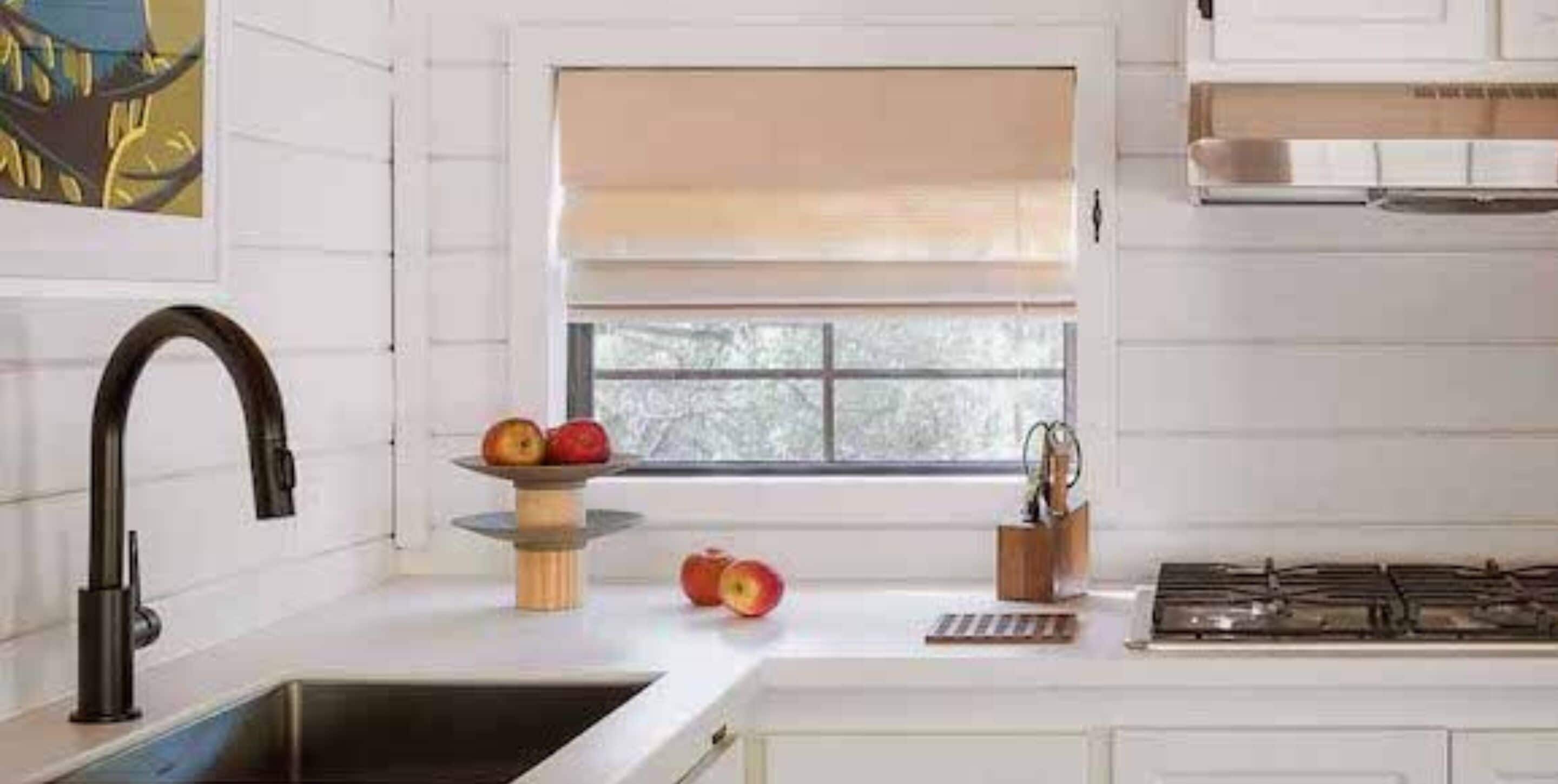 A kitchen with a modern faucet, a window with blinds, a fruit bowl with apples, and a stove.