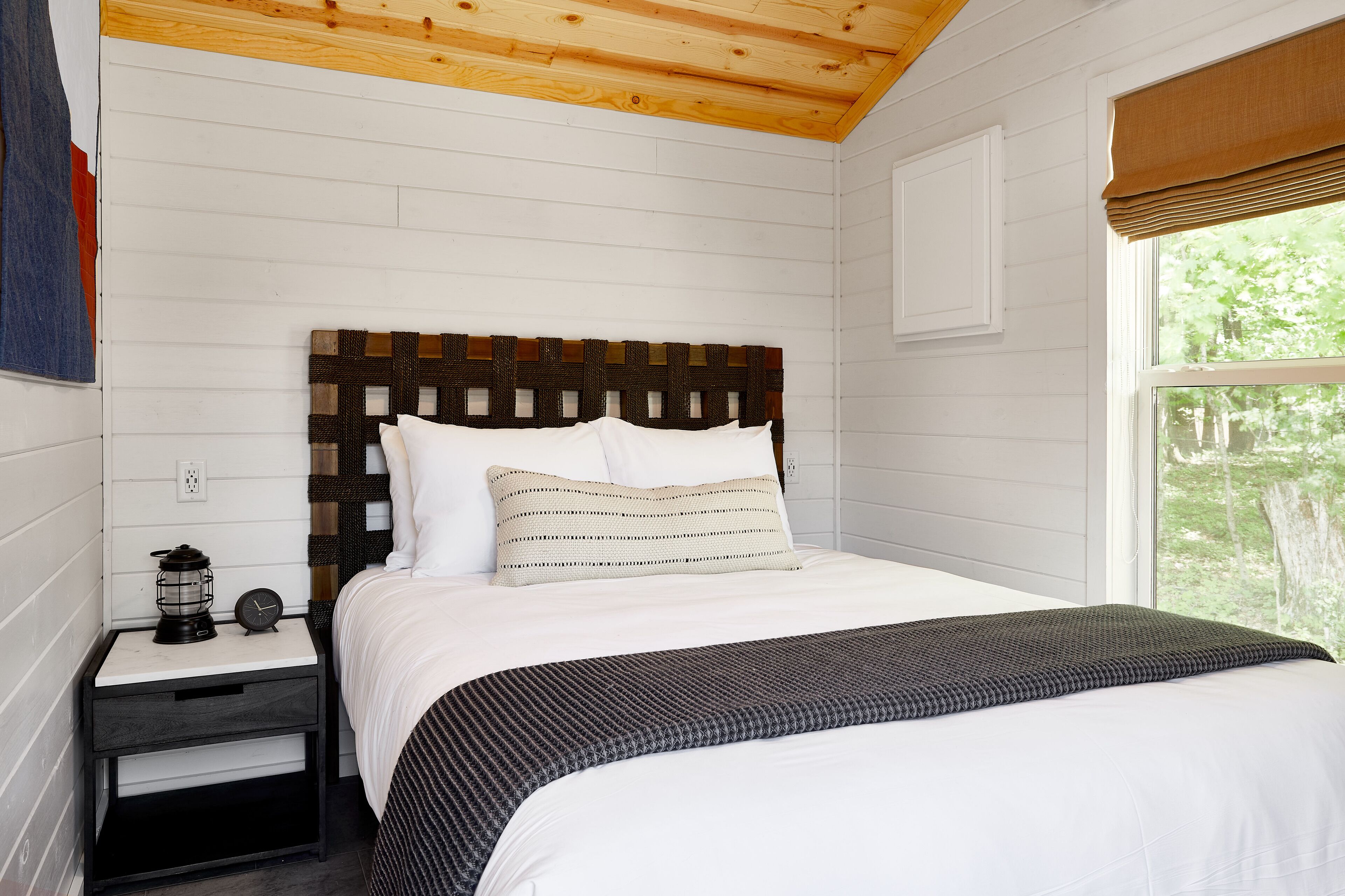 A neatly made bed with a patterned headboard, a bedside table with a lantern and clock, and a window with a view of trees.