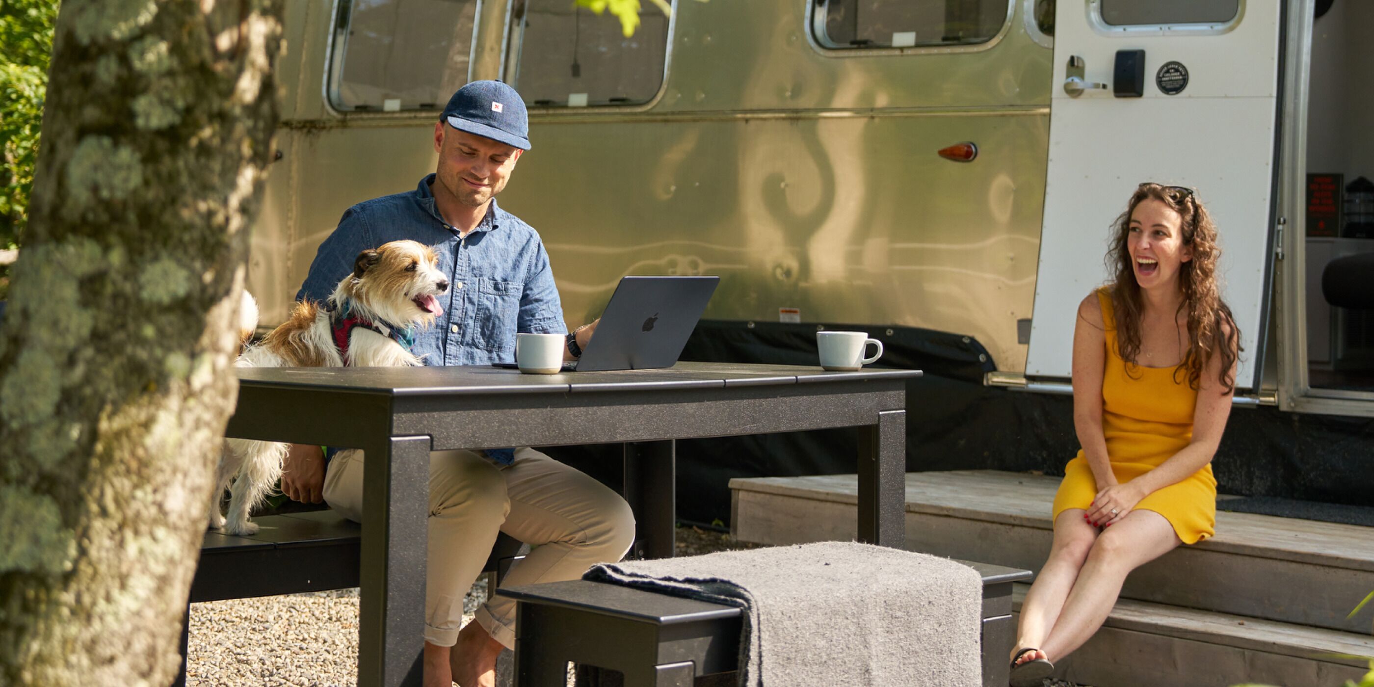 Un hombre y una mujer sentados en una mesa al aire libre con una laptop y una taza, un perro sobre la mesa, y una caravana de fondo.