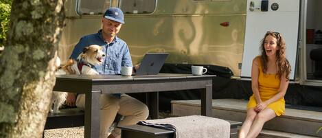 Un hombre y una mujer sentados en una mesa al aire libre con una laptop y una taza, un perro sobre la mesa, y una caravana de fondo.
