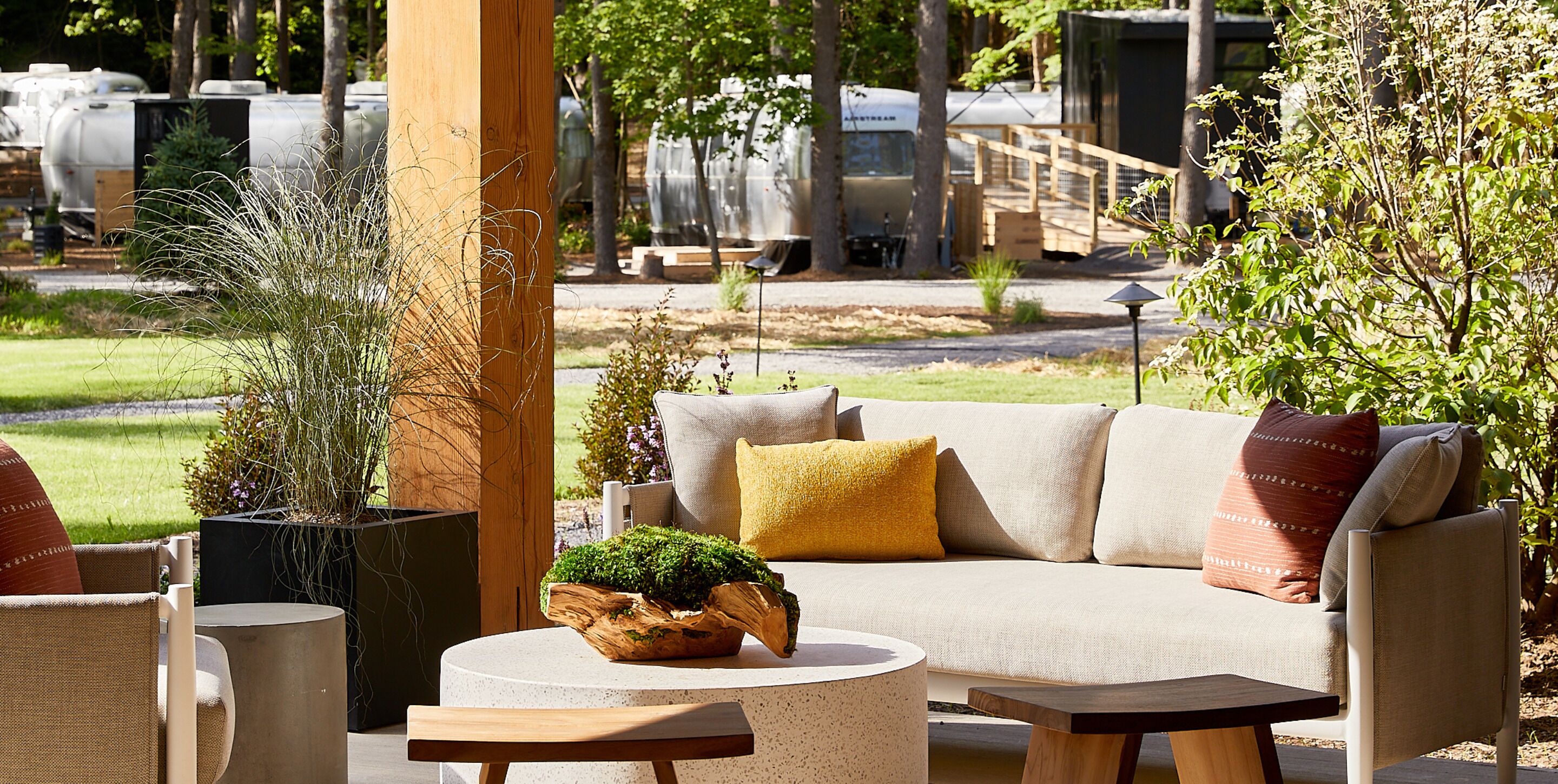 A cozy outdoor seating area with a sofa, chairs, and a round table, surrounded by potted plants and a wooden structure.