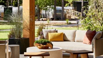 A cozy outdoor seating area with a sofa, chairs, and a round table, surrounded by potted plants and a wooden structure.