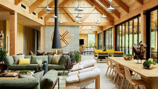 Amplia sala de estar con techo alto, vigas de madera y ventanas grandes. Hay varias disposiciones de asientos, incluyendo un sofá, sillas y una mesa de comedor.