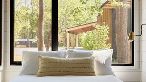 Una cama con sábanas blancas y un cojín tejido, una ventana con vistas a árboles y una estructura de madera, y una lámpara fijada a la pared.