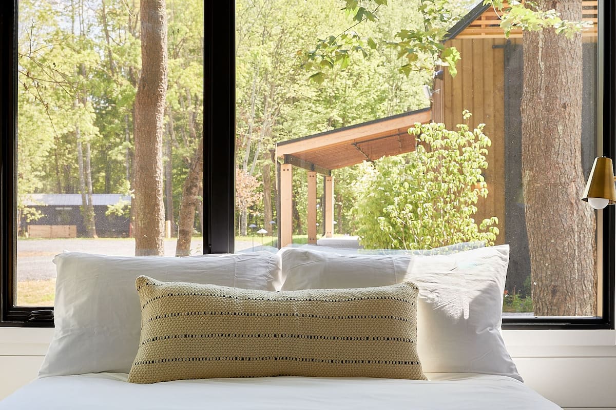 Una cama con sábanas blancas y un cojín tejido, una ventana con vistas a árboles y una estructura de madera, y una lámpara fijada a la pared.