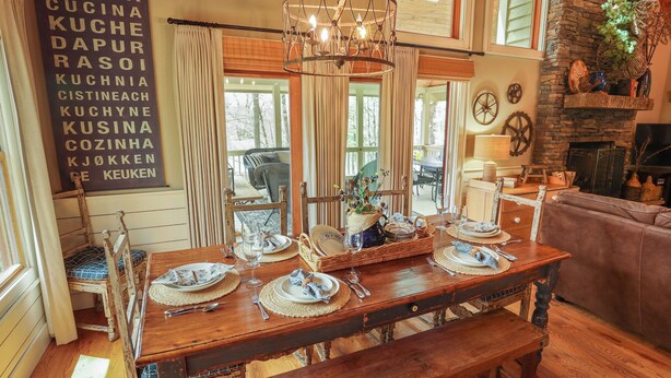 Un coin repas avec une table en bois dressée pour quatre personnes, un lustre et une vue sur l’extérieur grâce à de grandes fenêtres.