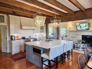 A kitchen with a large island, wooden beams, and a television above the fireplace.