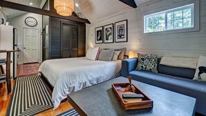 A bedroom with a bed, a sofa, a coffee table, a clock, and framed pictures on the wall. - Owl's Den: Easy Walk to Main Street | Adorable Tiny Home w/ Screened Porch (Highlands)