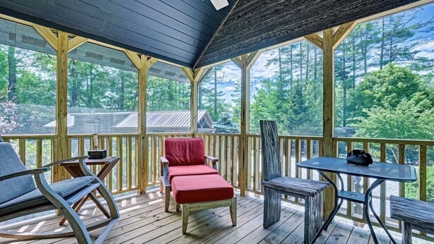 A covered deck with wooden railings, a rocking chair, a red bench, and a table with chairs. - Owl's Den: Easy Walk to Main Street | Adorable Tiny Home w/ Screened Porch (Highlands)