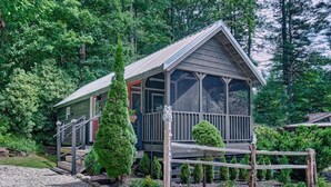 A wooden cabin with a porch, surrounded by dense forest. - Owl's Den: Easy Walk to Main Street | Adorable Tiny Home w/ Screened Porch (Highlands)