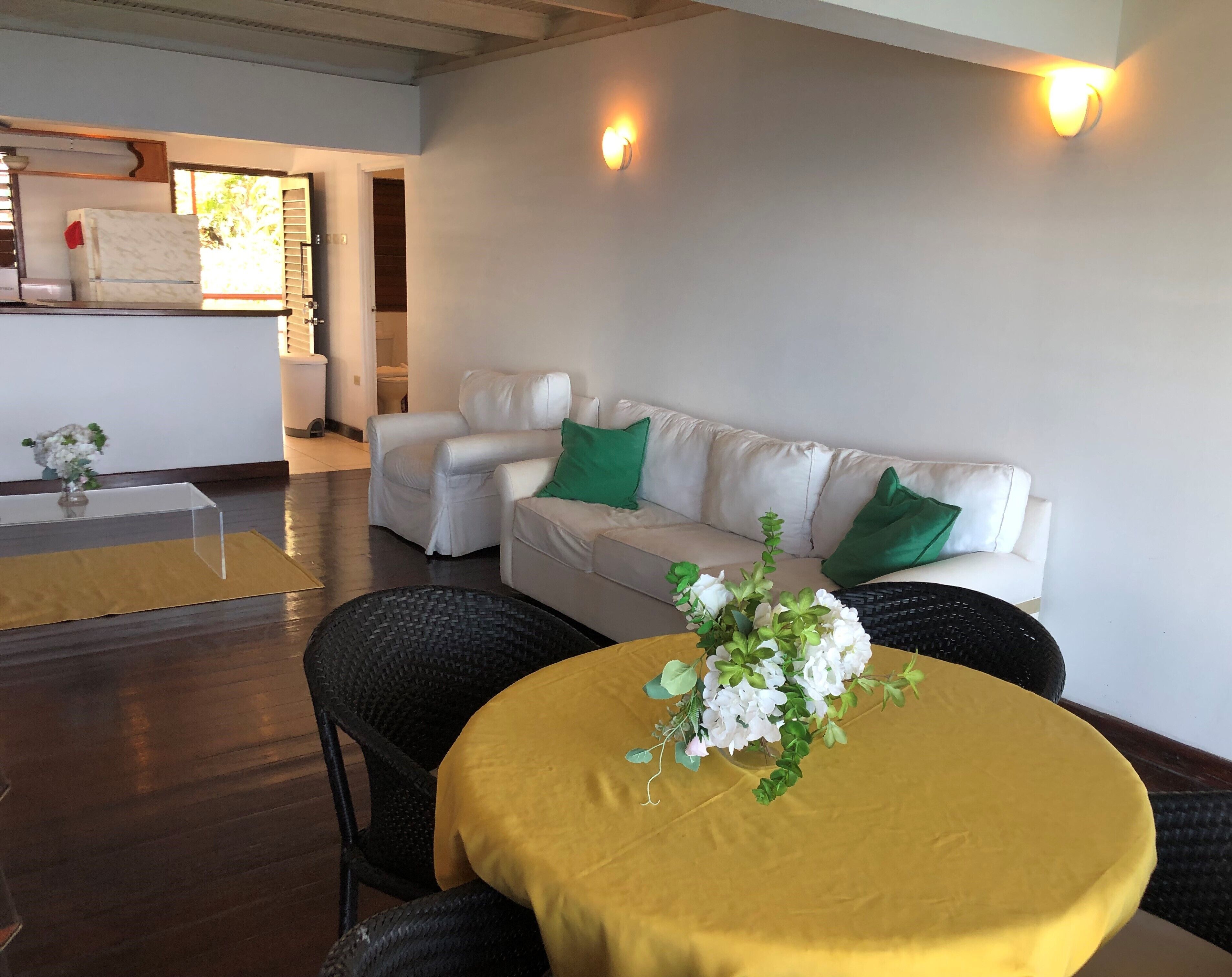A modern living room with a white sofa, green cushions, a round table with a yellow tablecloth, and a vase of flowers.