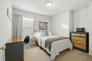 A bedroom with a bed, dresser, chair, bedside table, lamp, and a wall-mounted picture.