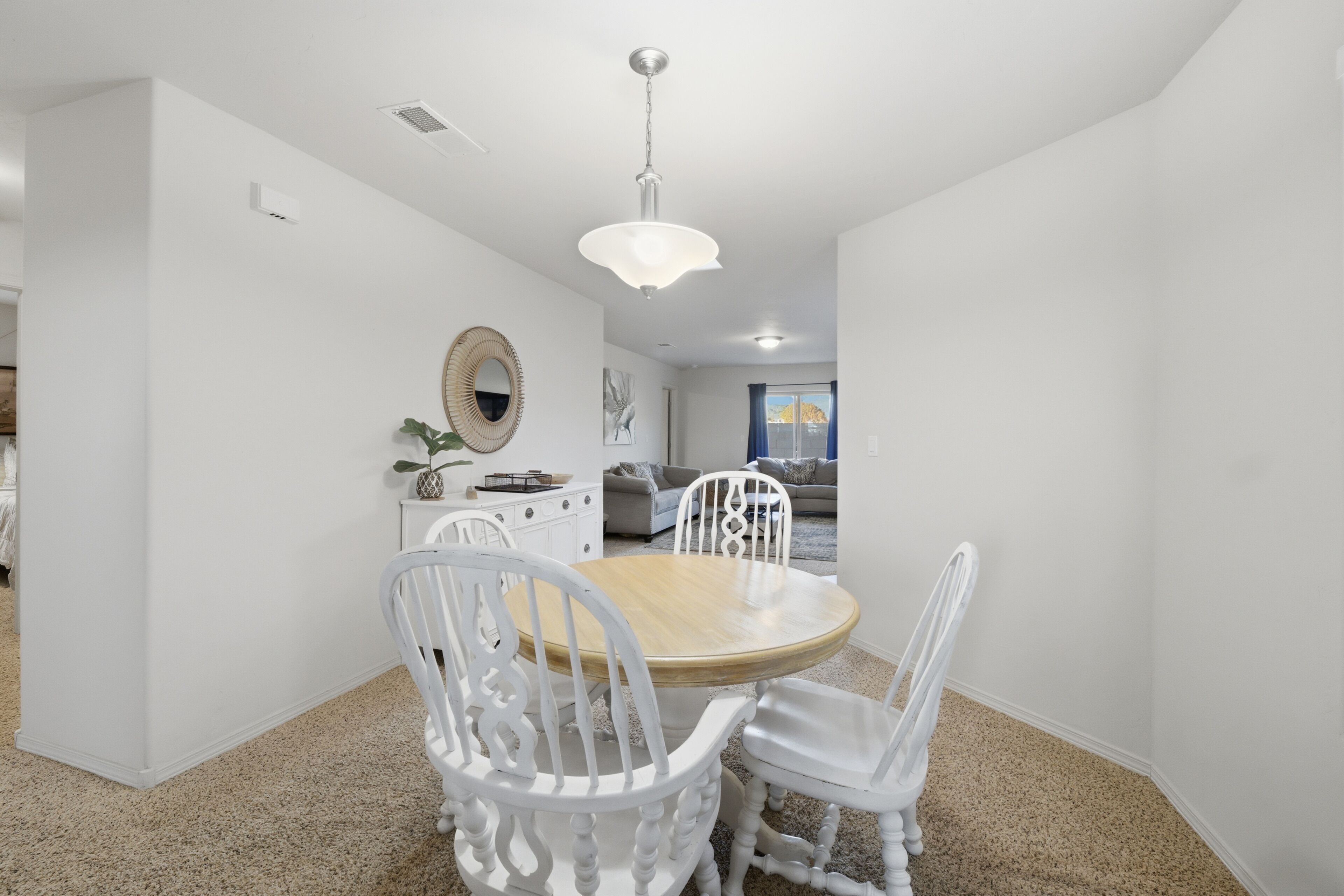 A dining area with a round wooden table and white chairs, a hanging light fixture, and a white wall with a decorative mirror.