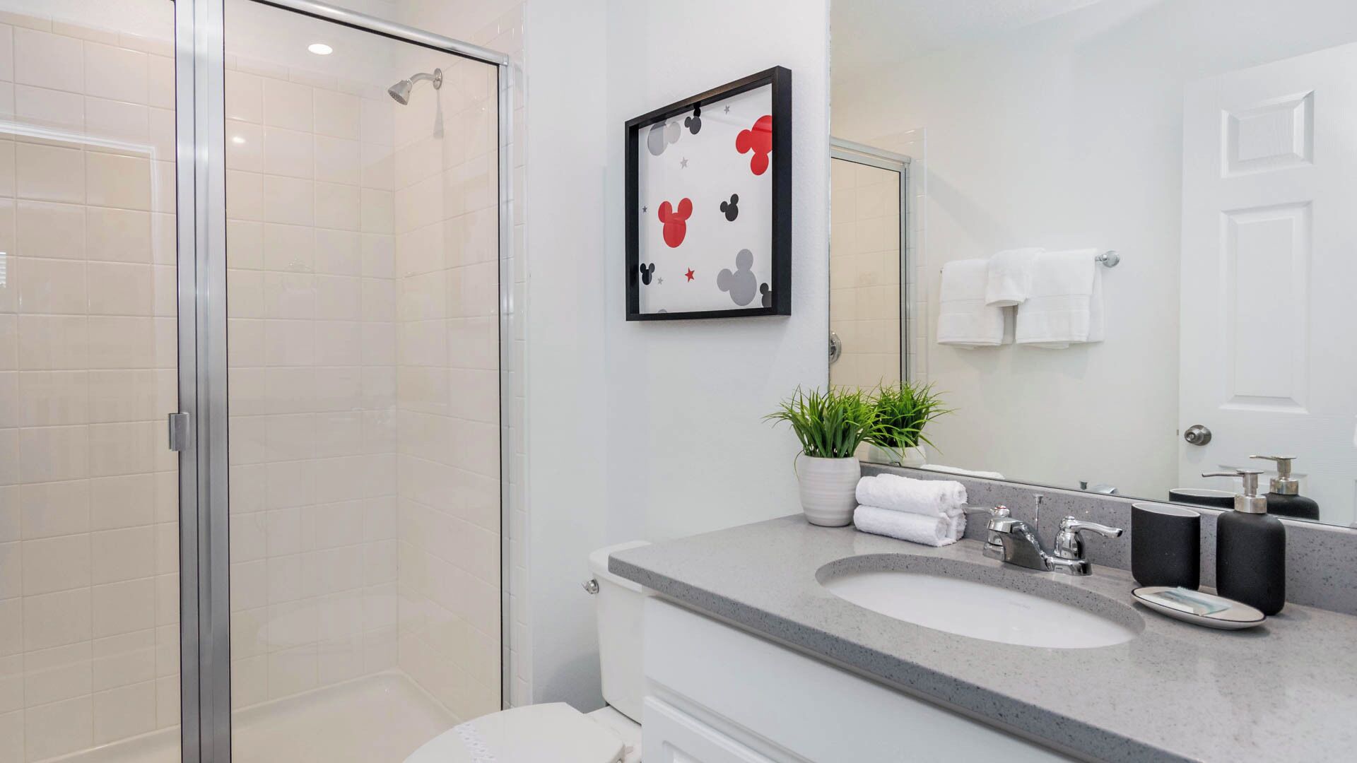 A modern bathroom with a glass shower enclosure, a large mirror, a framed Mickey Mouse picture, a potted plant, and a countertop with a sink and soap dispenser.