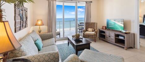 A living room with a sofa, chairs, a coffee table, a TV, and a view of the ocean through large sliding doors.