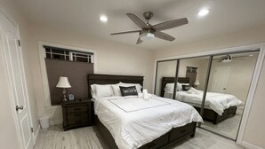 Une chambre à coucher avec un grand lit, une table de chevet, une lampe, un ventilateur de plafond, une fenêtre avec des stores et un grand miroir.