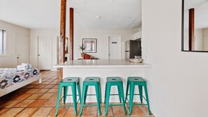 Eine moderne Küche mit einer zentralen Kochinsel, vier grünen Barhocker, einem Bett mit gemustertem Überwurf und einem großen Spiegel an der Wand.